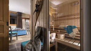 Una sauna de madera con bancos y un perchero para toallas, una silla con un abrigo de piel, y una ventana que muestra una habitación con esquíes y un sofá azul.