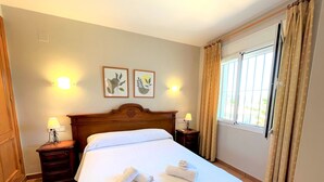 A neatly made bed with white linens and two rolled towels, a wooden headboard, bedside lamps, and two framed wall art pieces.