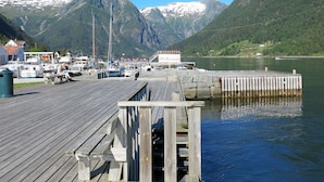 En träbrygga med förtöjda båtar, en liten byggnad med ett rött tak och berg i bakgrunden.