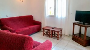 Uma sala de estar com um sofá vermelho, uma pequena mesa de madeira, uma televisão sobre um móvel e uma janela com cortinas.