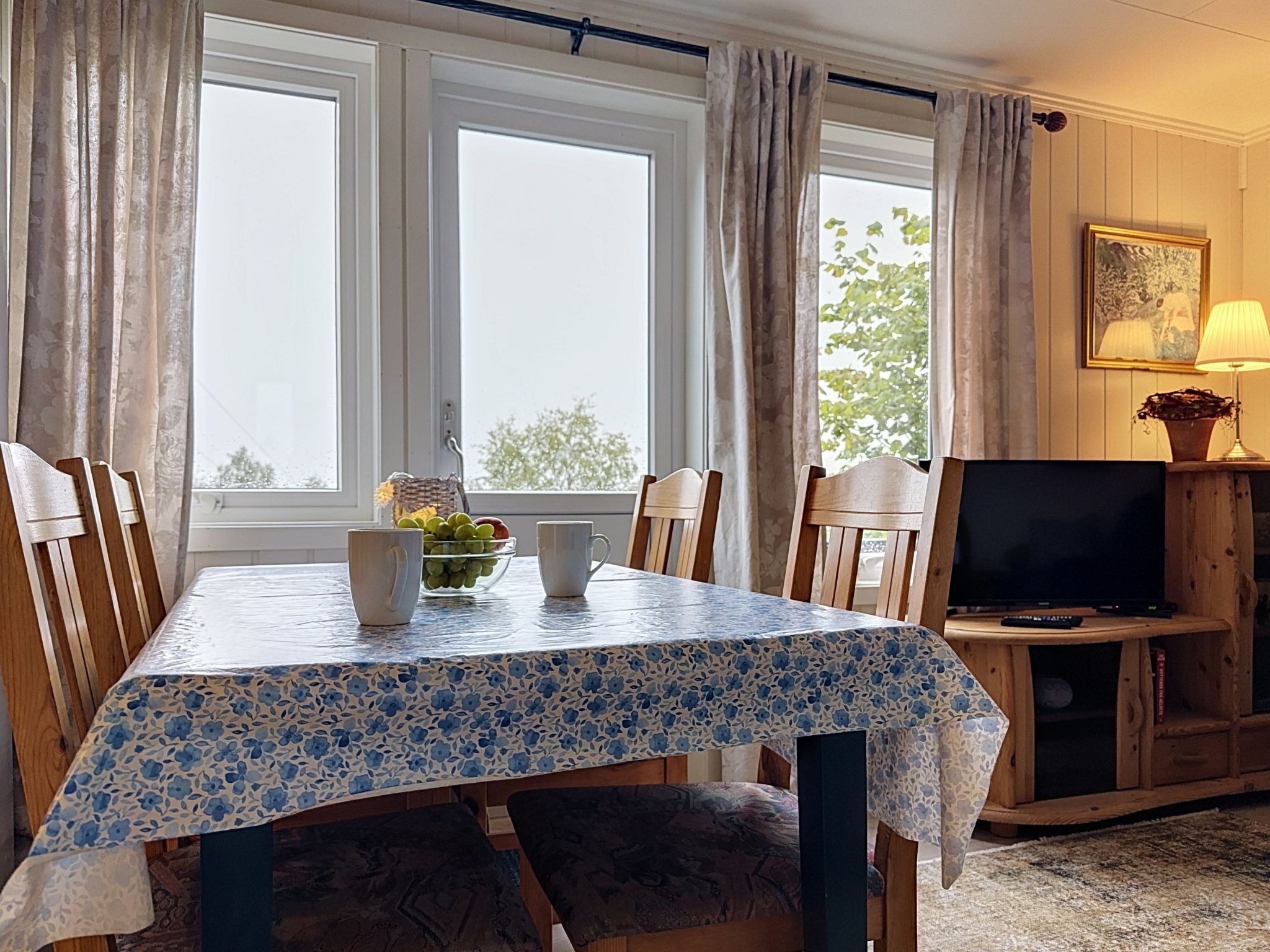 Ein Essbereich mit einem Tisch für zwei Personen, Stühlen, einem Fernseher und einem fenster mit Vorhängen.