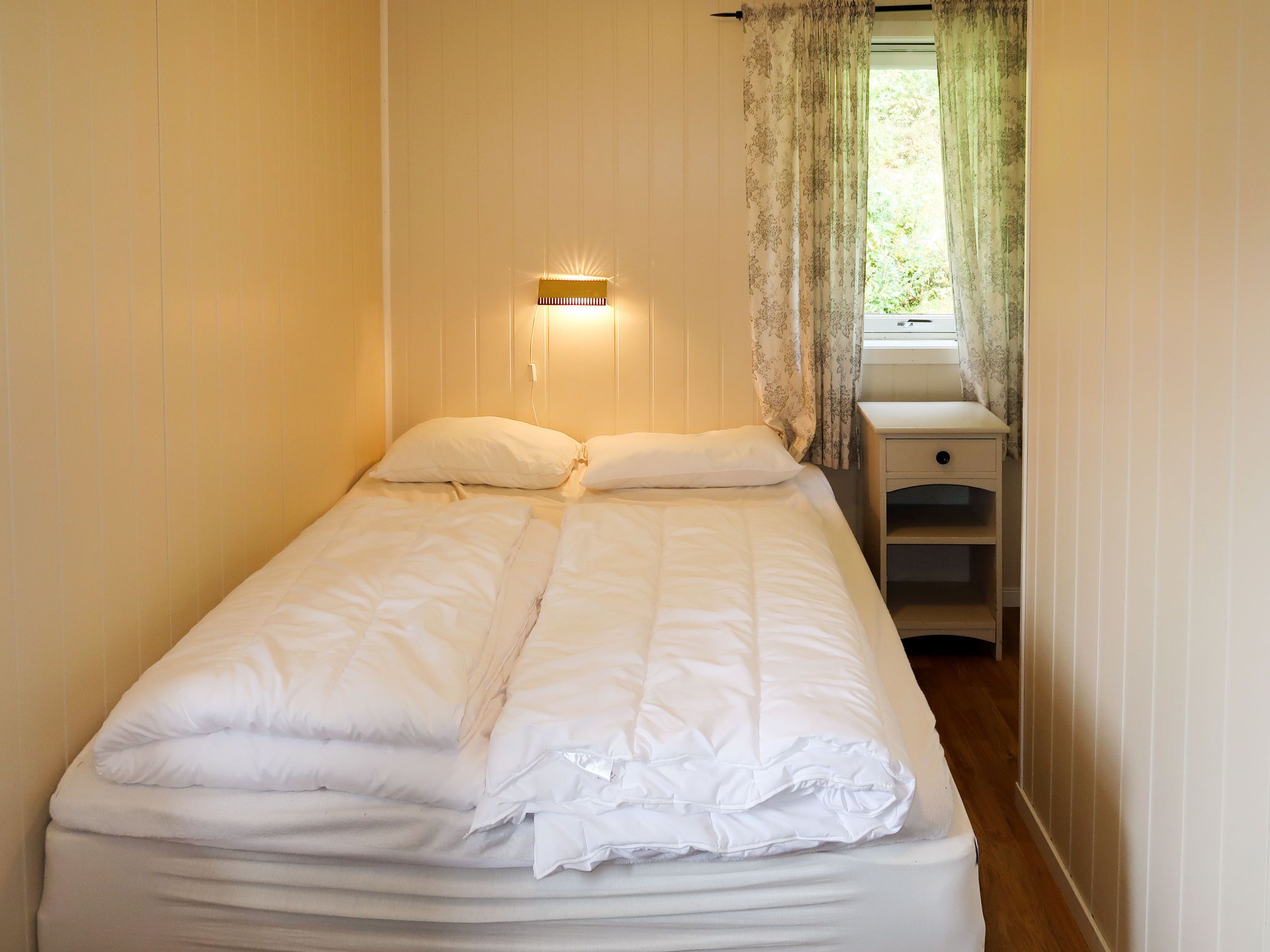A neatly made bed with white bedding in a room with wooden walls and a small bedside table with drawers.