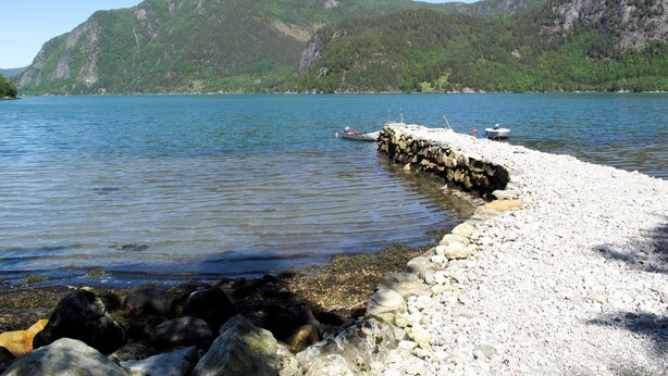 Un rivage rocheux avec une jetée qui s’avance dans des eaux calmes, le tout entouré de collines verdoyantes.
