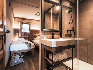 Ett kompakt hotellrum med träinredning, en stor spegel, ett handfat och en dusch. Det finns två sängar och ett litet bord med handdukar.