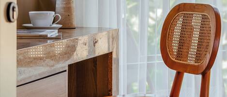 A wooden desk with a lamp, a cup, and a chair by a window with sheer curtains.