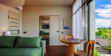 Una habitación de hotel moderna con un sofá verde, una mesa de comedor de madera y vistas a la ciudad a través de amplios ventanales.