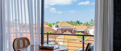 A balcony with a wooden table and chairs, a glass of water, and a view of buildings and greenery.