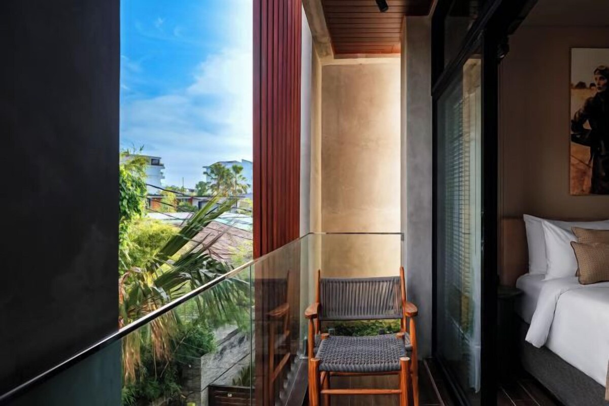 A balcony with a wooden chair, a bed with white bedding, and a view of greenery and buildings.