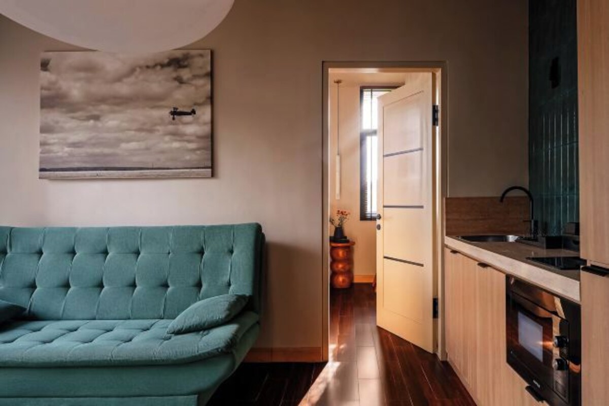 A modern living room with a teal sofa, a kitchen with wooden cabinets, and a painting of clouds on the wall.