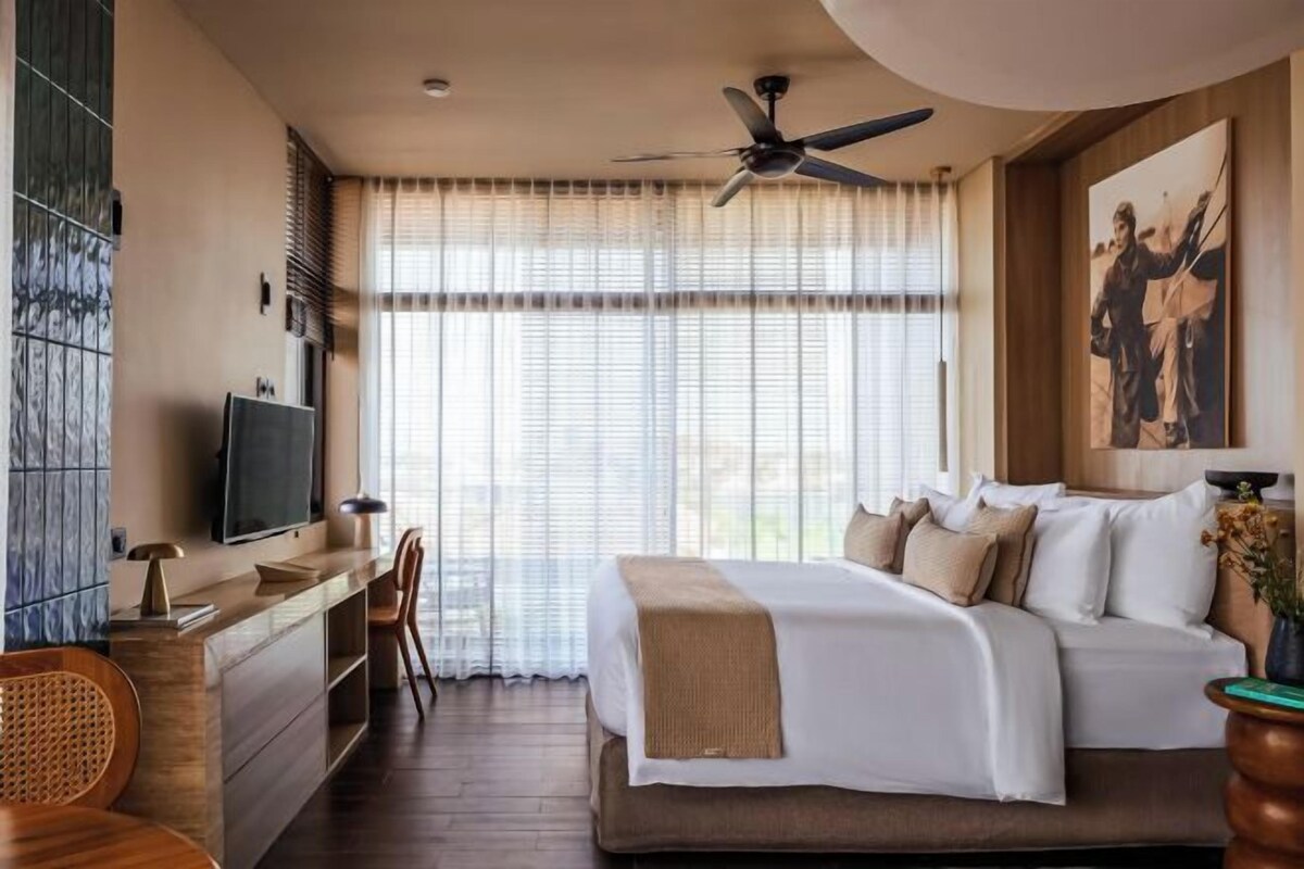 Una habitación de hotel moderna con una cama grande, un escritorio con televisor de pantalla plana, un ventilador de techo y ventanas amplias con cortinas ligeras.