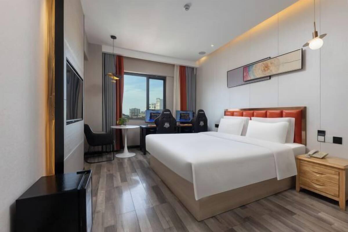 A hotel room with a large bed, a desk with two chairs, a small table, a nightstand, and a window with a city view.