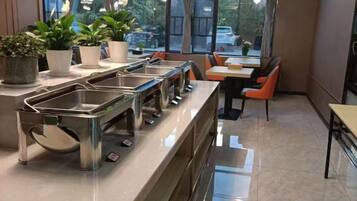 A buffet setup with multiple stainless steel serving stations, potted plants, and seating arrangements.