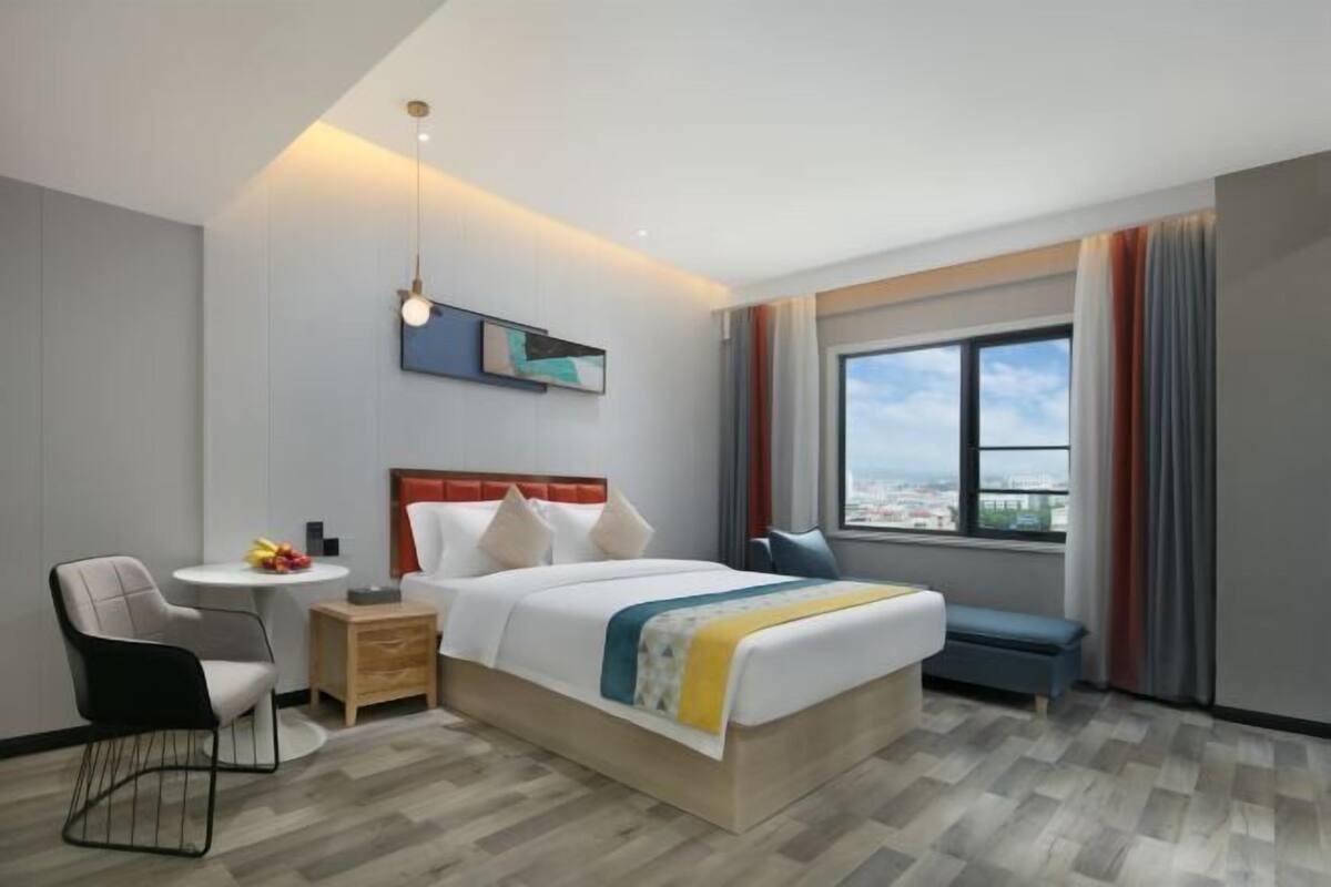 A hotel room with a bed, bedside table, chair, and a window with a city view.