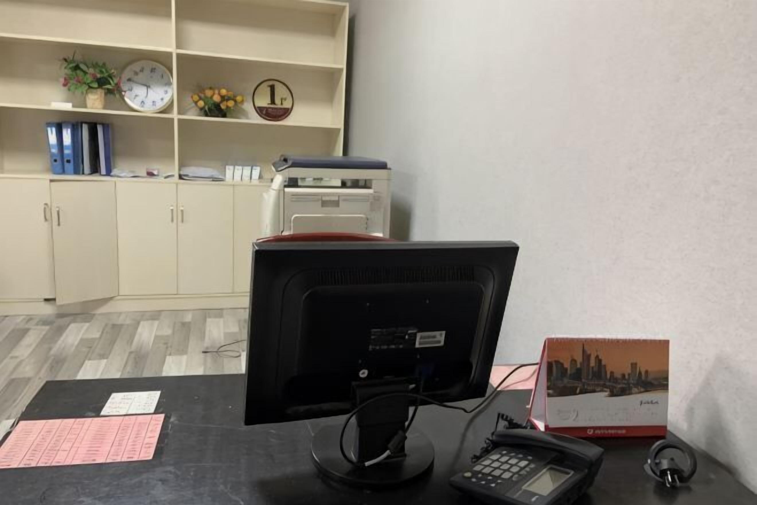 A desk with a computer monitor, keyboard, and telephone. There is a clock, a plant, and a shelf with binders and a printer.