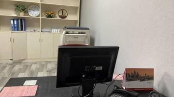 A desk with a computer monitor, keyboard, and telephone. There is a clock, a plant, and a shelf with binders and a printer.