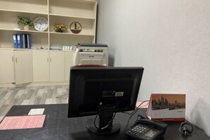 A desk with a computer monitor, keyboard, and telephone. There is a clock, a plant, and a shelf with binders and a printer.