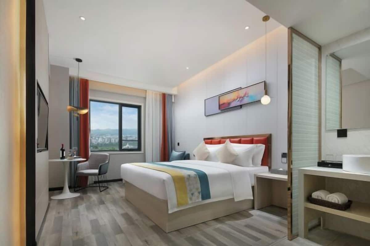 A modern hotel room with a large bed, a desk, a chair, a small table, a window with a view, and a bathroom with a mirror and shelves.