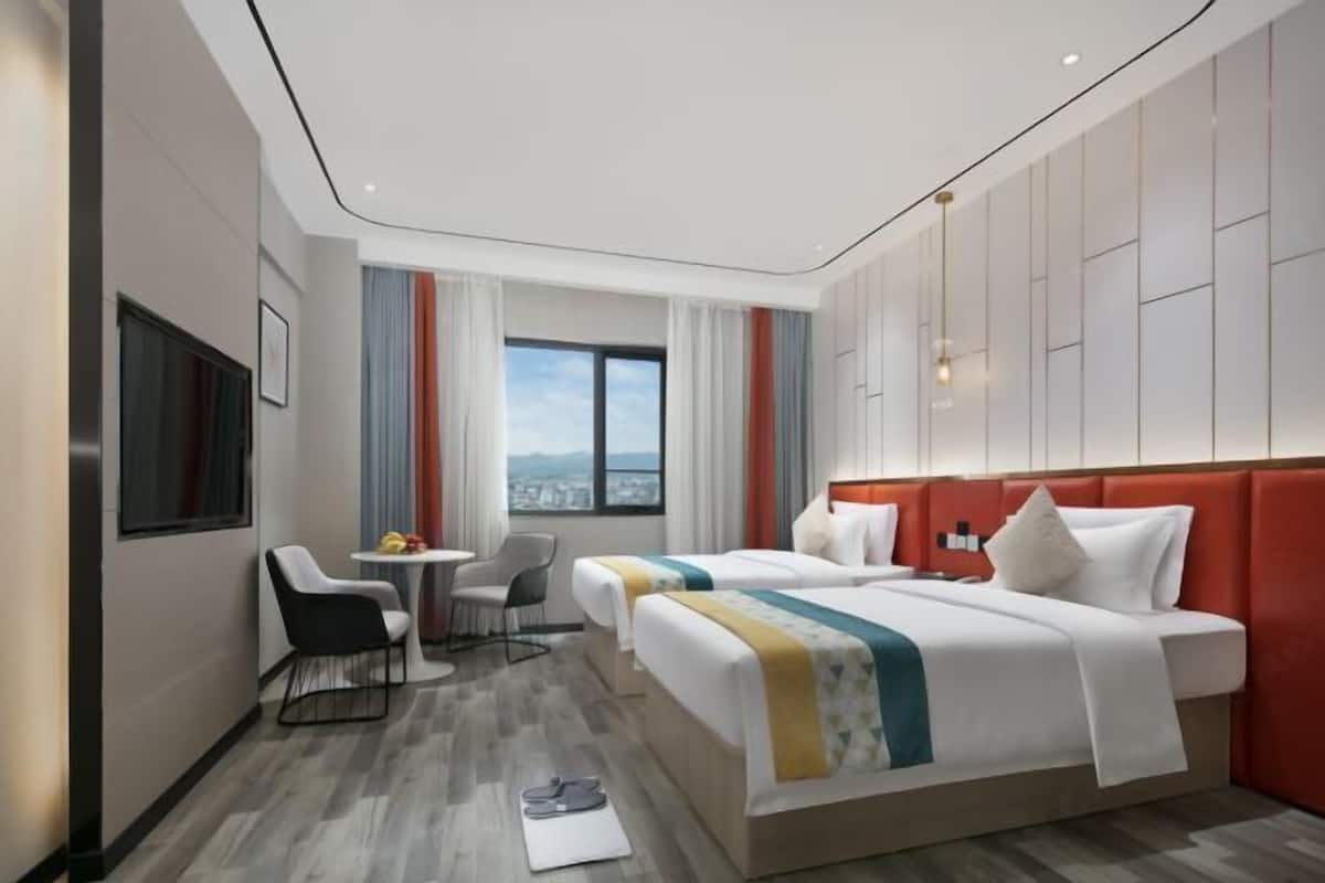 A hotel room with two beds, a desk, a chair, and a window with a city view.