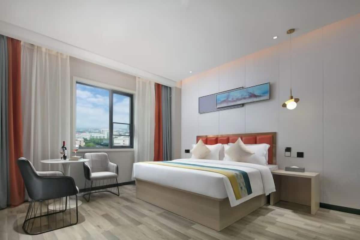 A modern hotel room with a large bed, a small table, two chairs, a nightstand, a painting, and a window with a city view.