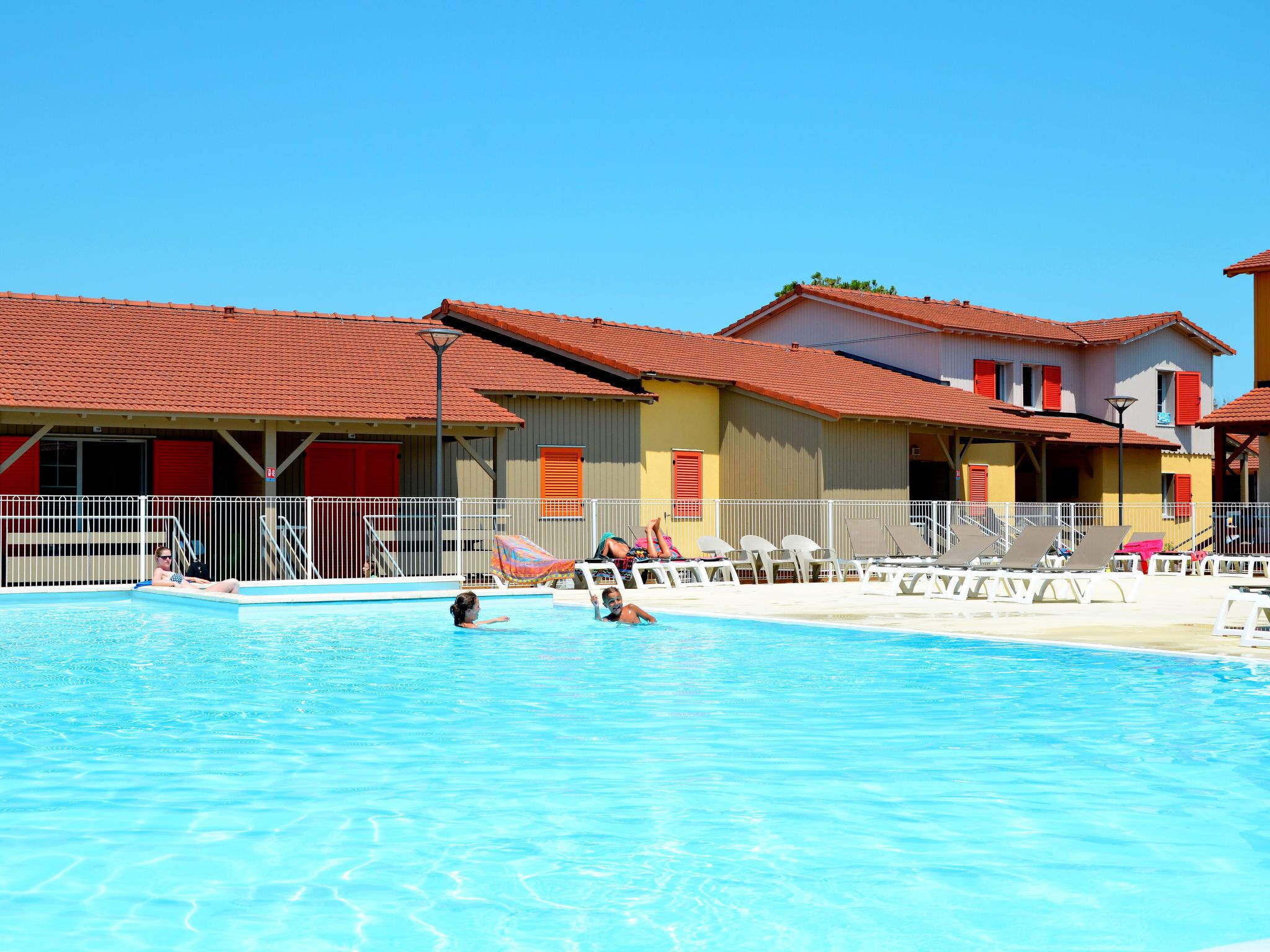 En swimmingpool med solstolar och en byggnad med röda tak och vita räcken.