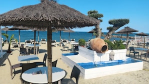 Een terras met tafels en stoelen, een grote terracotta pot en een fonteintje op een zandstrand met parasols en palmbomen.
