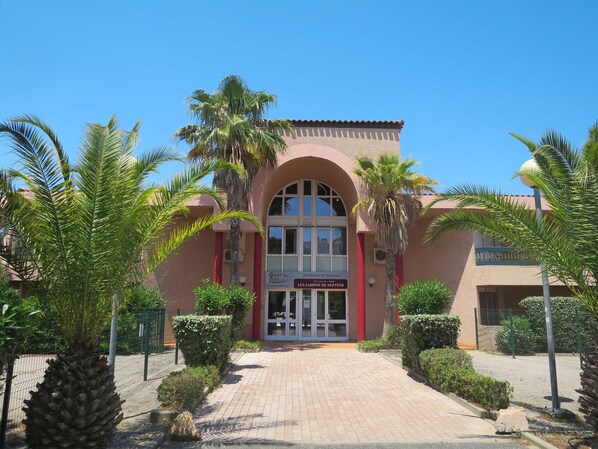 A building with a red facade, arched windows, and a sign reading "Grand Bleu Residences de Tourisme Les Jardins de Neptune".