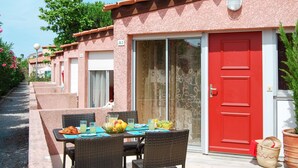Fila de edificios rosados con números en las puertas, una puerta roja, y una mesa con sillas y fruta.