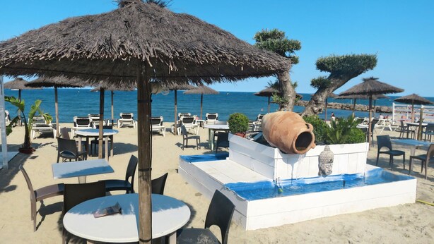Un área con mesas y sillas al aire libre, una maceta de barro grande y una pequeña fuente de agua en una playa arenosa con sombrillas y palmeras.