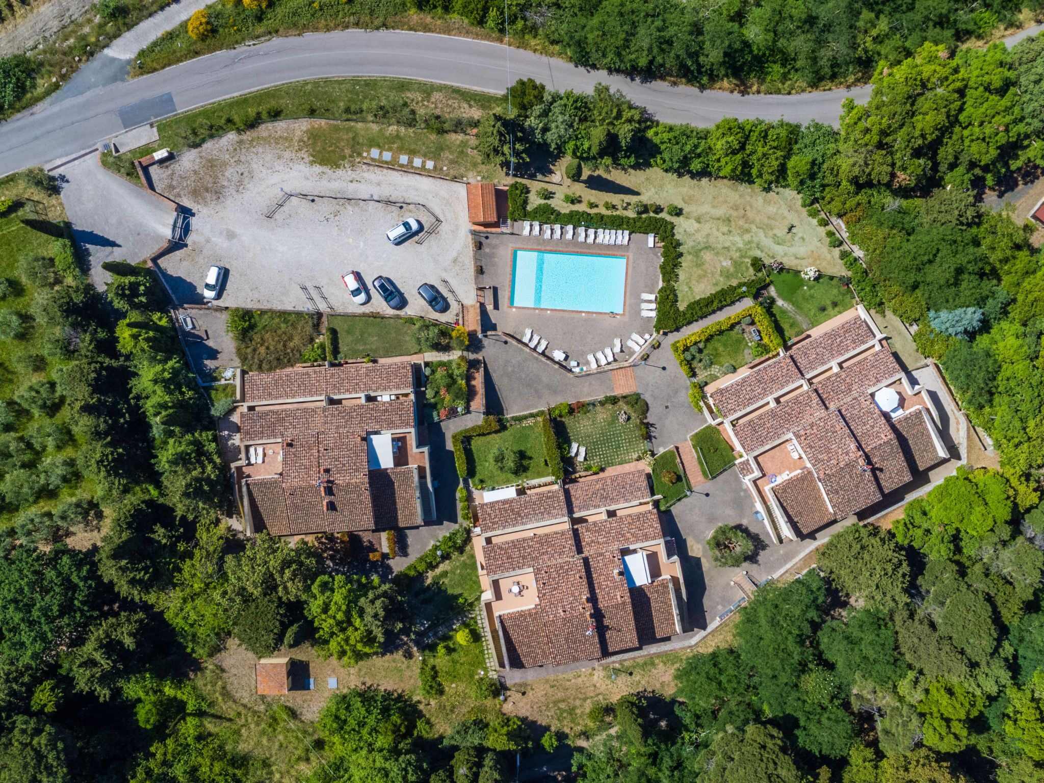An aerial view of a residential area with multiple terracotta-roofed houses, a swimming pool, and a parking lot.