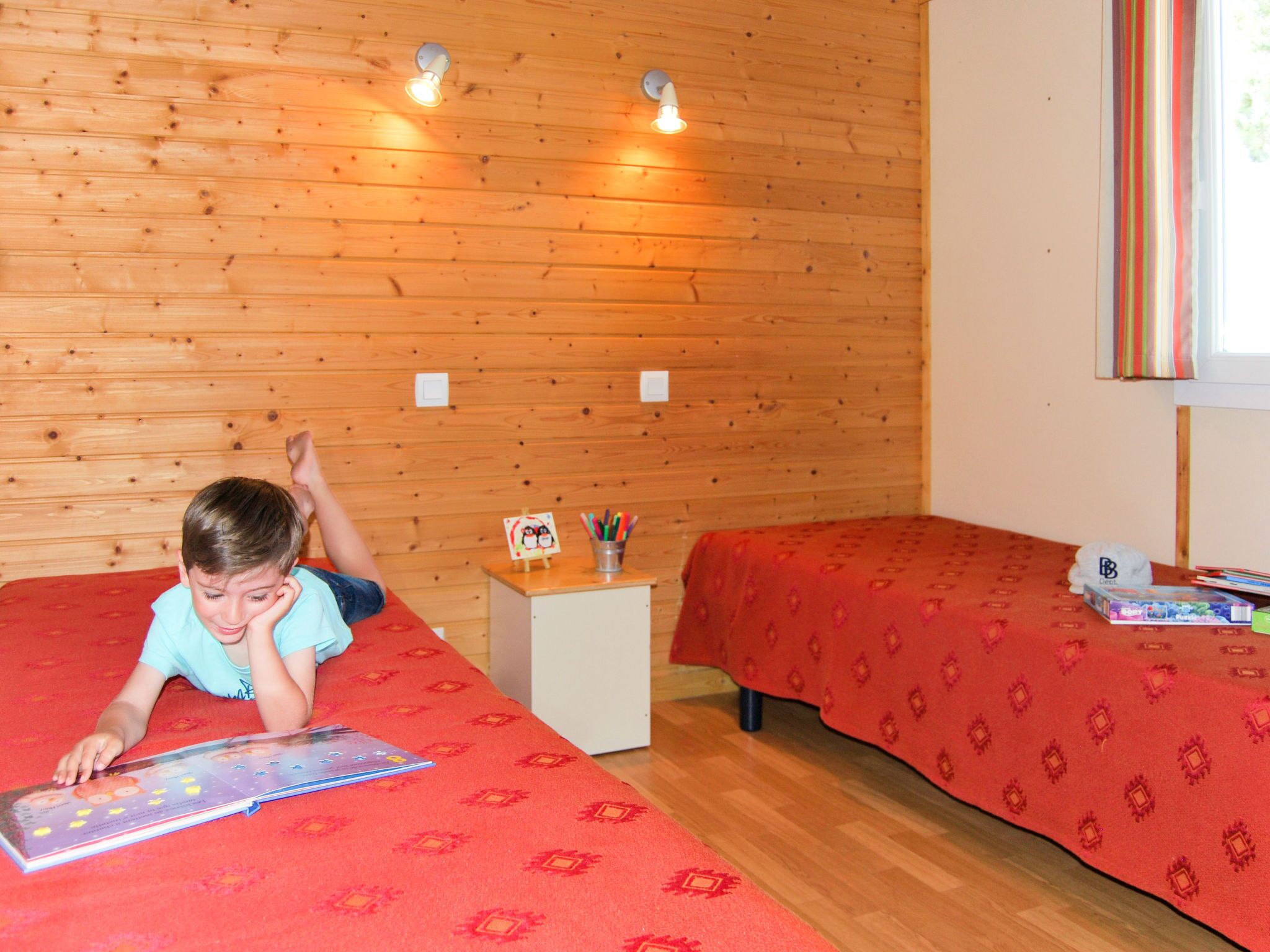 Une pièce aux murs en bois, avec deux lits recouverts de draps rouges, une petite table sur laquelle se trouve un cadre photo et des crayons de couleur, et une fenêtre ornée d’un rideau à rayures.