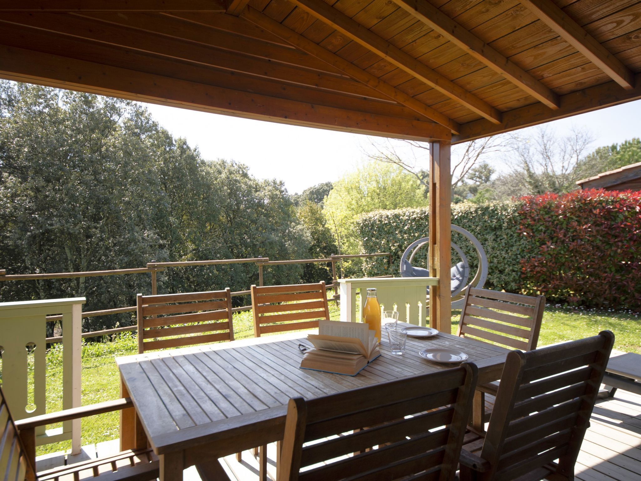 An outdoor wooden deck with a table set for two, surrounded by chairs, overlooking a garden with trees and shrubs.