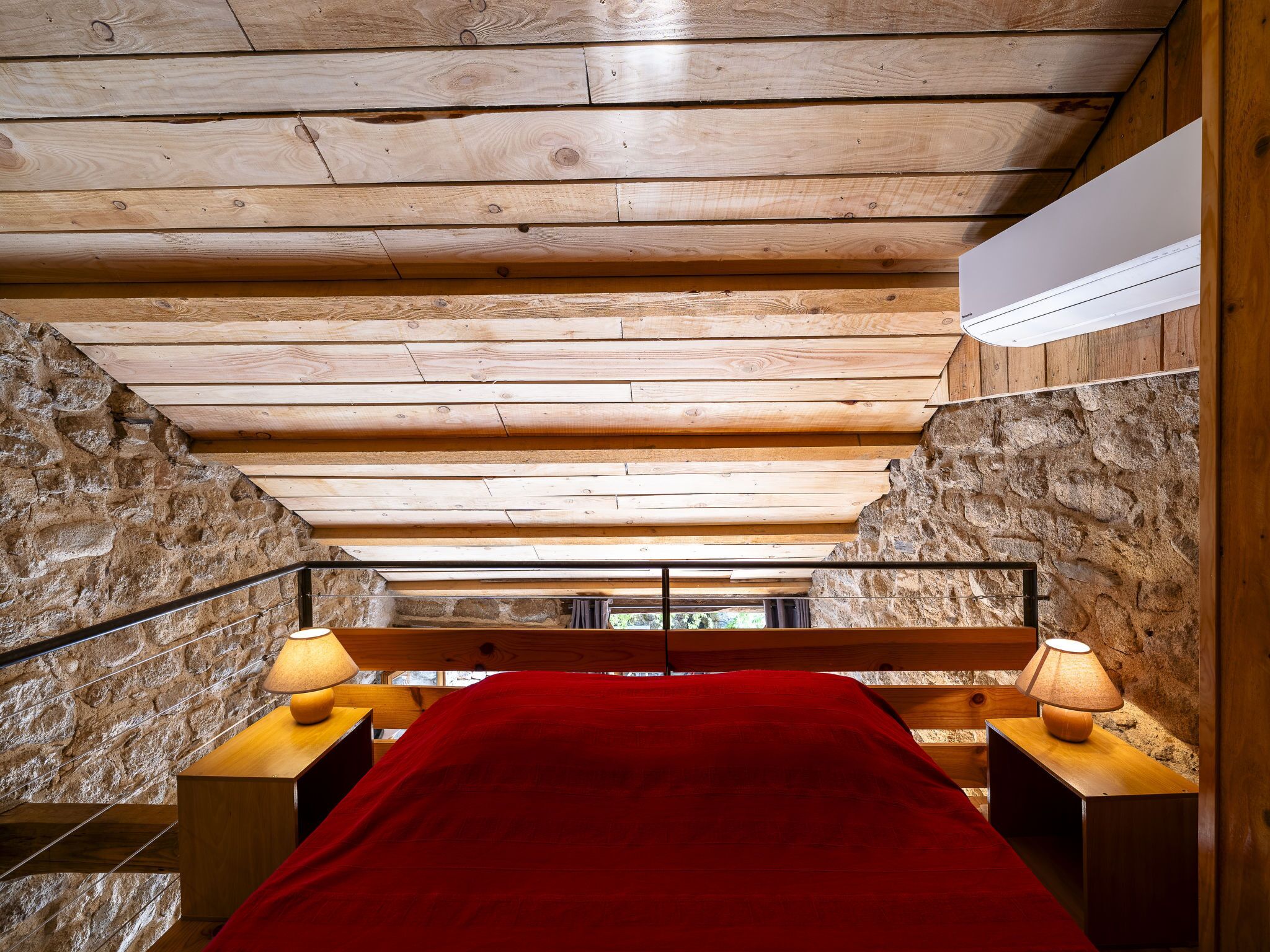 Ein Schlafzimmer mit Holzboden, Steinwänden, einem Bett mit roter Decke und zwei Nachttischlampen.