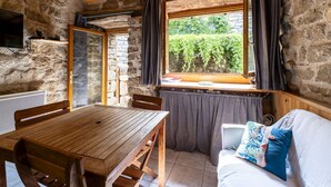 Ein gemütlicher Raum mit Holztisch und Bänken, einer Steinsäule, einem Fenster mit Vorhängen und Blick ins Grüne.