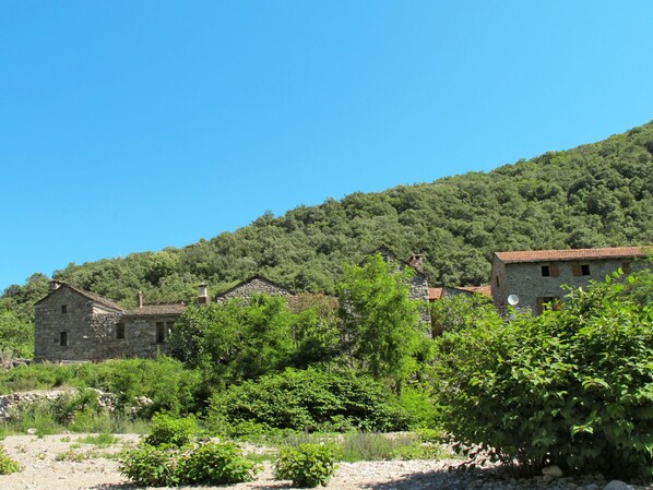 A stone building with a tiled roof, surrounded by lush greenery and a hillside in the background. - Chassezac by Interhome (Les Vans)