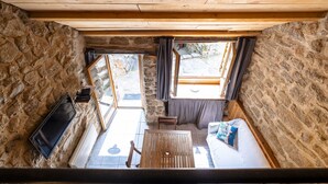 Ein rustikaler Raum mit Steinwänden und Holzbalkendecke, einem Fernseher, einer Couch mit Kissen, einem Tisch und einem Fenster mit Vorhängen.