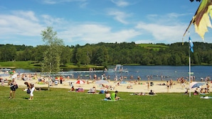Una playa con gente tomando el sol y nadando, un área verde y un lago al fondo.