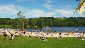 A beach with people sunbathing and swimming, a grassy area, and a lake in the background.