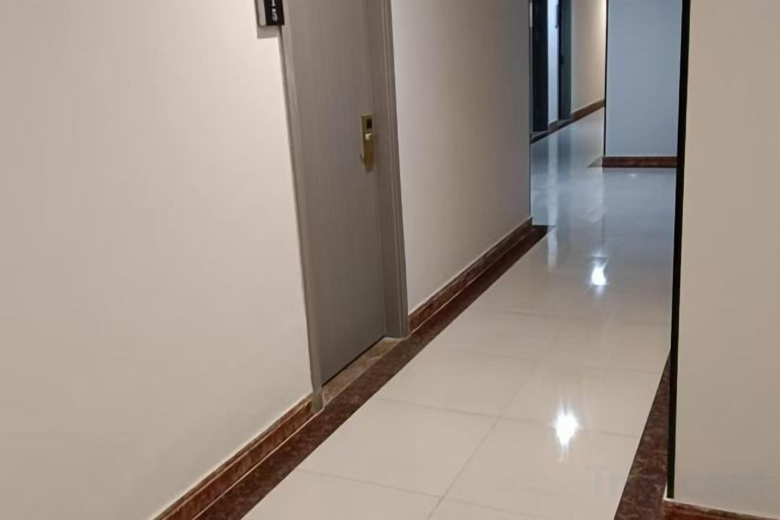 A corridor with a door on the left, a reflective tiled floor, and a plain wall.