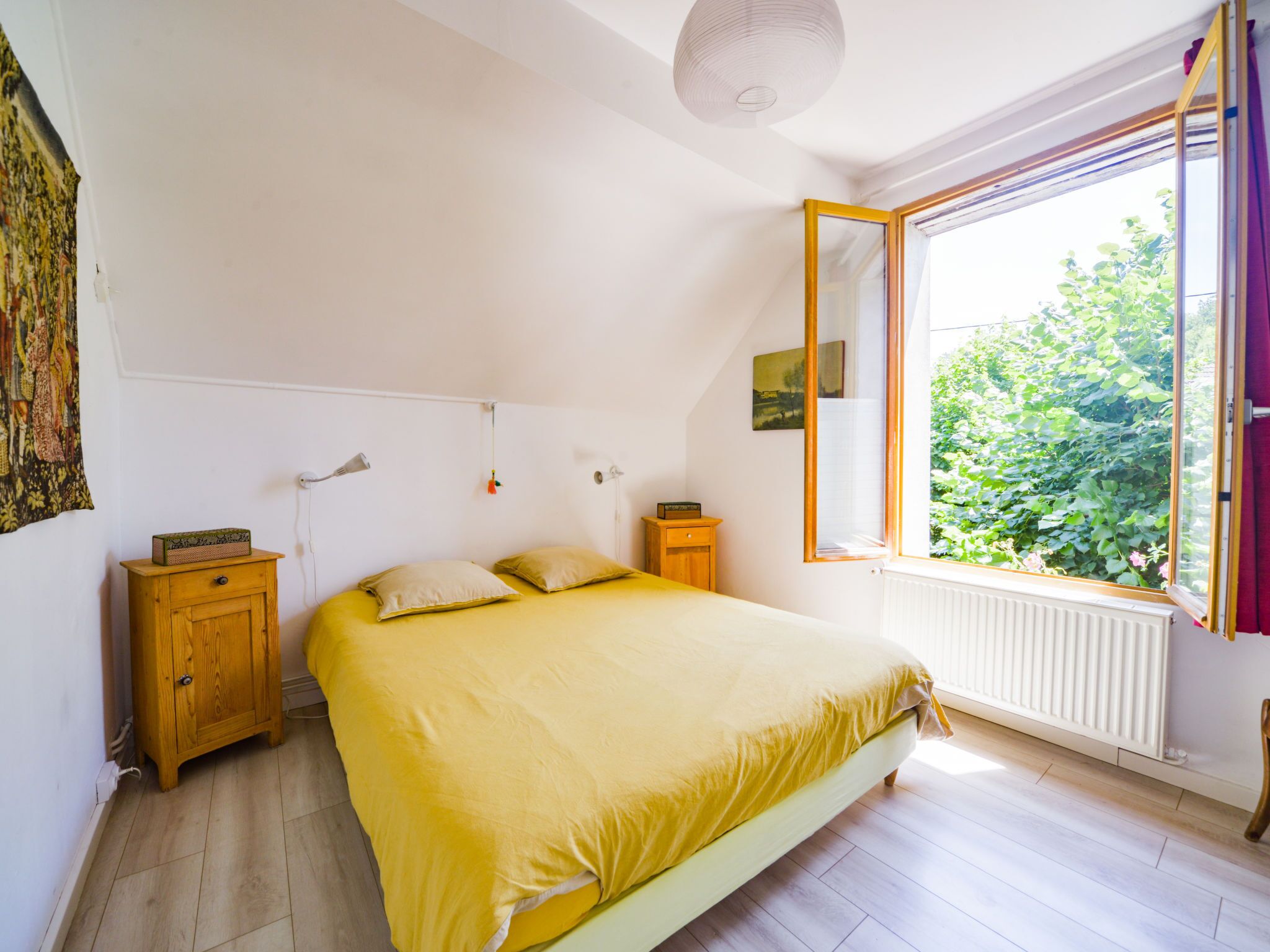 A bedroom with a bed, a wooden nightstand, a radiator, a window with a view of greenery, and a ceiling light.