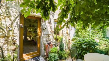 Un bâtiment en pierre avec une porte en bois, un petit espace jardin avec une chaise et une table, et une plante en pot.