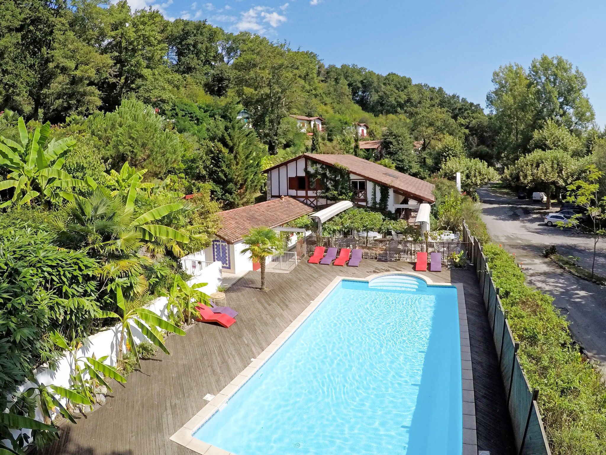 A swimming pool with lounge chairs, surrounded by lush greenery and a building with a red roof.
