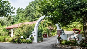 Una entrada con puerta metálica y un arco blanco, rodeada de abundante vegetación y un edificio con techo rojo.