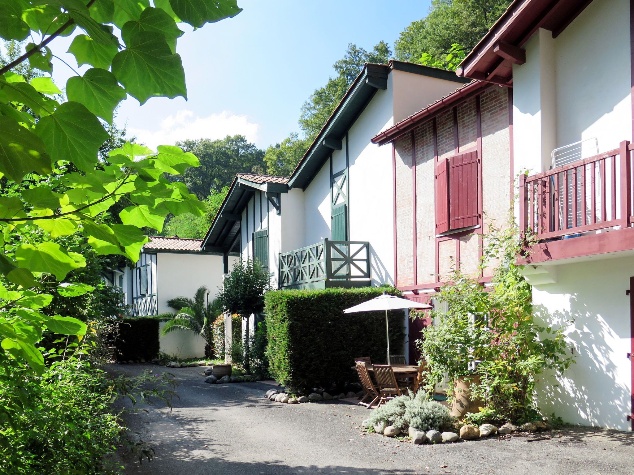 A narrow driveway leading to a row of traditional houses with wooden shutters and a small patio area with outdoor furniture.