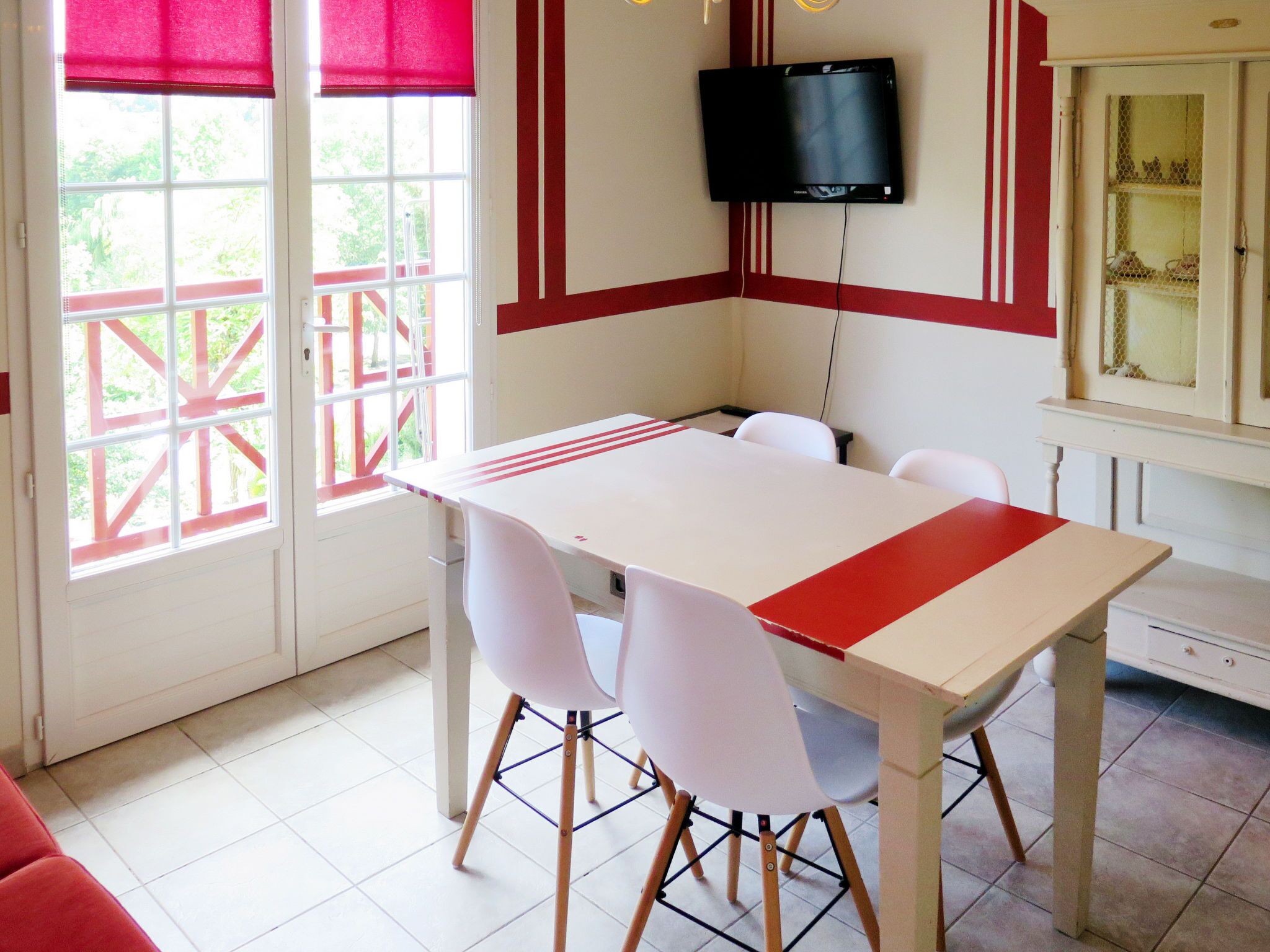 A dining area with a white table and chairs, a television mounted on the wall, and a window with red blinds.