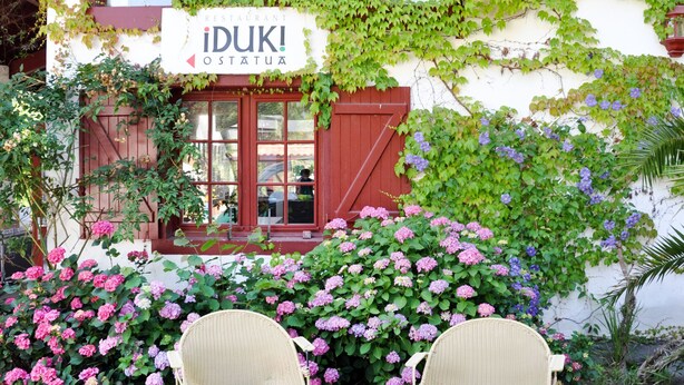 Un restaurante llamado 'iDUK! OSTATUA' con puerta roja y ventanas, rodeado de exuberante vegetación y arbustos de hortensia rosa.