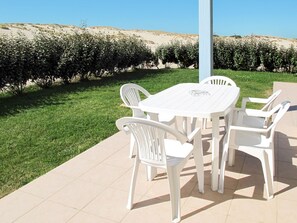 Eine Terrasse im Freien mit weißem Tisch und Stühlen, umgeben von einem gepflegten Rasen und einem Heckenstreifen.