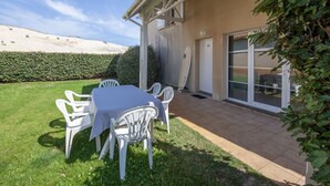 A patio with a table and chairs, a surfboard leaning against a wall, and a view of a beach in the background. - Résidence Dune Blanche - Océan1 by Interhome (Biscarrosse)