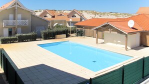 A swimming pool surrounded by buildings with red-tiled roofs, a clear sky, and a hilly landscape in the background. - Résidence Dune Blanche - Soleil by Interhome (Biscarrosse)