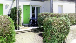 A building with a green door, a small patio with a table and chairs, and a hedge in the foreground. - Les Terrasses du Lac by Interhome (Mimizan)