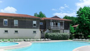 A swimming pool with lounge chairs, a wooden building with a balcony, and a clear blue sky. - Les Terrasses du Lac by Interhome (Mimizan)
